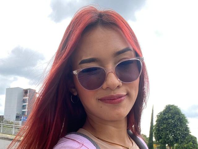 SashaHans, avec des cheveux roux, portant des lunettes de soleil et une chemise rose, souriant avec un petit piercing au nez et des boucles d'oreilles pendantes, se tient debout à l'extérieur sous un ciel nuageux..