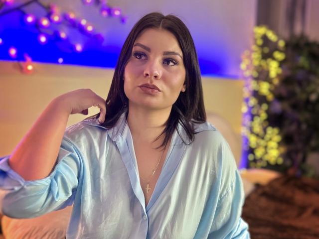 AmandaHoppe has medium-length black hair and light skin. She wears a shiny, light blue satin button-up shirt with the collar open, exposing a bit of her chest. Her right hand is raised, touching her hair. She has a neutral expression, looking slightly to the left. A subtle gold necklace is visible. The background is blurred with blue and yellow lights, and a plant is on the right. The image has a soft, warm light.