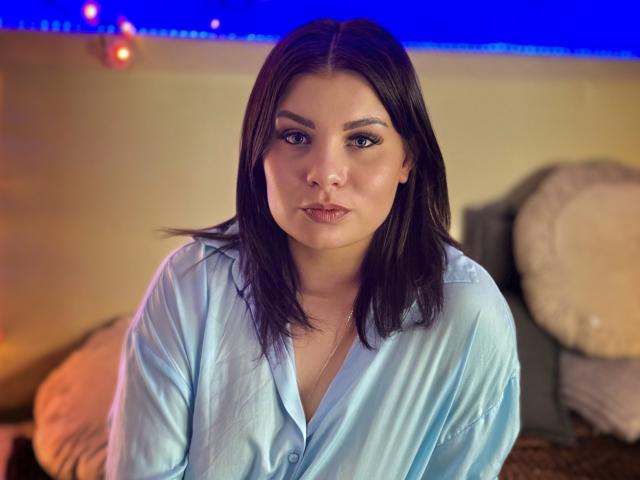 AmandaHoppe is in a close-up photograph. She has medium-length, straight black hair and is wearing a light blue button-up shirt. Her facial expression is neutral with slightly parted lips. She has dark, defined eyebrows, and her makeup includes dark eyeliner. The background is blurred, with a blue strip of LED lights and circular, beige pillows. The lighting is warm, with a small orange light on the left. The overall composition is centered on AmandaHoppe, who looks directly at the camera.