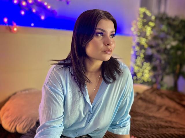 AmandaHoppe with dark brown hair, wearing a light blue shirt, pouting her lips, looking to the side, on a brown bed, with a plant in the blurred background.