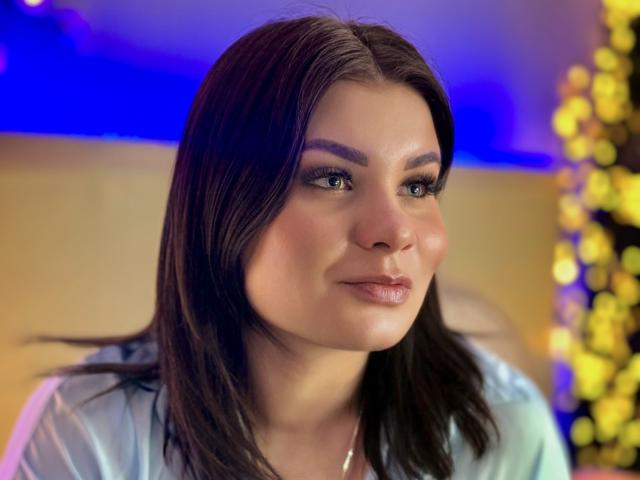 AmandaHoppe has a fair skin tone and dark brown, shoulder-length hair. She has green eyes with heavy, dark eyeliner and long, voluminous eyelashes. Her lips are full and glossed with a subtle pinkish tint. She wears a light blue shirt. The background is blurry with blue and yellow lights. Her expression is calm and slightly smiling. The photo is close-up, focusing on her face. She has a slight blush on her cheeks. The lighting highlights her facial features.