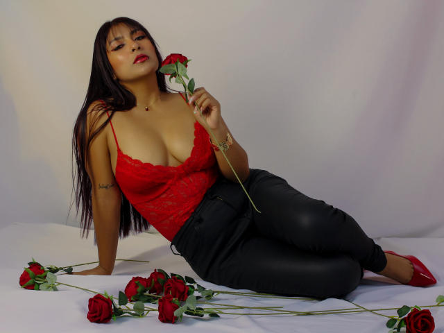 EvelynJohnsoon with long black hair, red lace top, black leather pants, red heels, holding rose, lying on bed, surrounded by roses, showing big tits, red lips, tattooed arm.