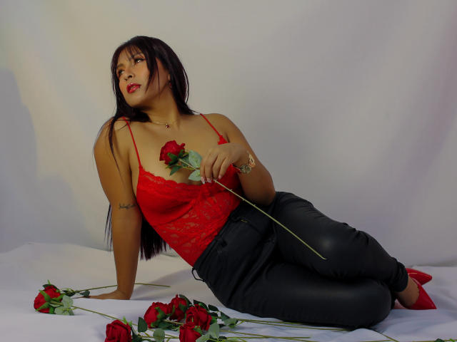 EvelynJohnsoon with long black hair, red lace top, black pants, holding rose, laying on white, surrounded by red roses, wearing red heels, tattoo on right arm.