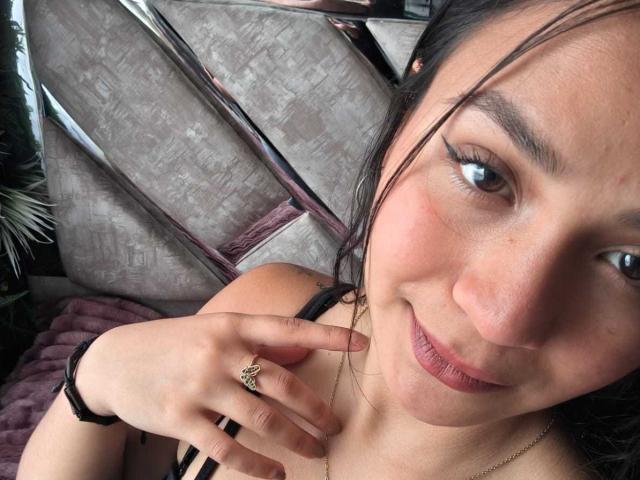 RouxiMori with dark hair, light skin, and brown eyes, smiling, wearing a black strap, gold ring, and bracelet, hand near face, subtle makeup, slightly messy.