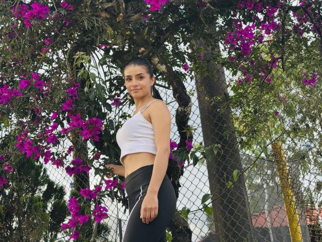 KimmyK with medium tits, white crop top, black pants, standing against a tree with purple flowers, black hair in a ponytail, looking at camera, outdoors, daytime, relaxed.