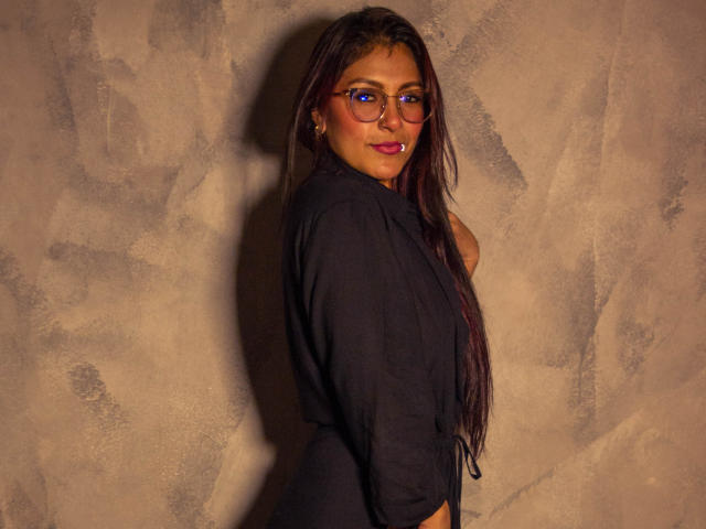 EvelynJohnsoon with long dark hair, glasses, and red lipstick, wearing a black shirt, showing her tits, against a textured wall, looking over her shoulder, smiling.