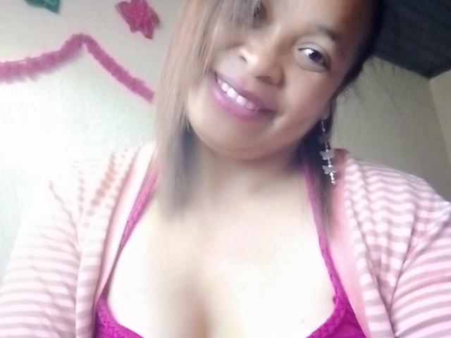 Rahel69 with medium brown skin, smiling, pink-striped cardigan, magenta lace bra, small tits, pink lipstick, brown hair, cross earrings, standing in soft light, looking at camera.