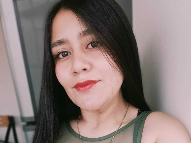 AnaVolkova with long black hair, small nose ring, red lipstick, green sheer top, smiling, brown eyes, light skin, wearing a thin necklace, in a selfie.