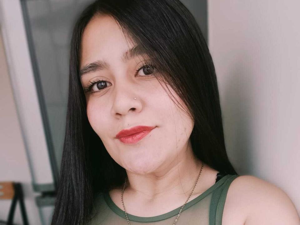 AnaVolkova with long black hair, small nose ring, red lipstick, green sheer top, smiling, brown eyes, light skin, wearing a thin necklace, in a selfie.