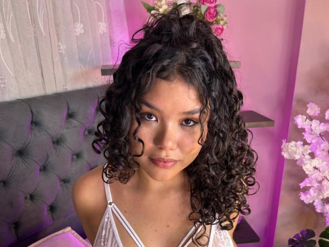 AnniePleasure has curly black hair in a high ponytail, looking directly at the camera with a neutral expression. She has dark brown eyes, full lips with light pink lipstick, and a medium skin tone. She wears a white, strappy top. The background includes a grey tufted headboard, pink walls, and floral arrangements. The lighting is soft, with purple and pink hues. Her makeup is subtle, emphasizing her natural features.