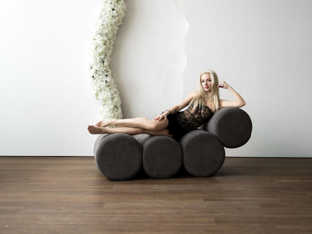 LexyGlam is a blonde woman with long hair, reclining on a dark gray, modern chaise made of circular, plush cushions. She wears a black lace lingerie bodysuit, showcasing her slender, toned physique. Her right arm is bent, with her hand resting on her head, while her left arm rests on the cushion. She has a relaxed, confident expression. The background features a white wall with a vertical, textured floral arrangement on the left. The floor is wooden, contrasting with the minimalist, modern aesthetic of the chaise. The image has a clean, contemporary style with a focus on simplicity and elegance.