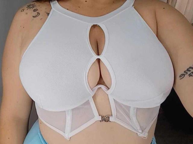 ValentinaDiazHot wearing a white cut-out top with large, oval-shaped openings over her tits, revealing a bit of cleavage. She has tattoos on her left shoulder and right arm.