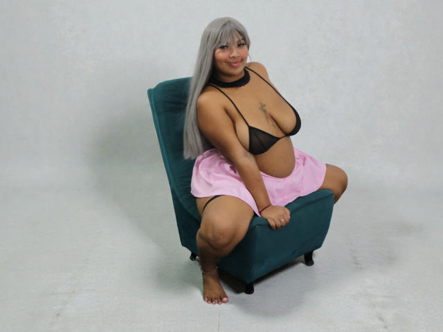 A photograph of DakoTaxt, a plus-sized woman with long, straight, gray hair and light brown skin, sitting on a teal upholstered chair. She wears a black mesh bra that exposes her large breasts and a pink skirt, with her legs spread apart. She has a small star tattoo on her chest and a black choker around her neck. She is barefoot, with toes painted pink. The background is plain and white.