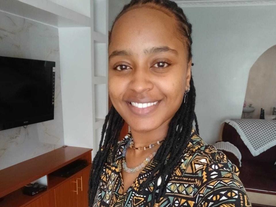 Close-up of Syrrenne with dark brown skin, smiling at the camera. She has thin, straight black braids pulled back. She wears a patterned shirt with geometric designs in yellow, black, and white, and a necklace with small beads. Her eyes are brown, and she has a small, even smile. The background shows a wall-mounted TV on the left and a bed with a checkered blanket on the right. The room has a mix of white and beige walls. Syrrenne looks relaxed and happy.