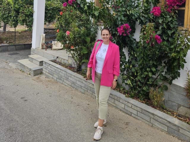CurvySweetGirl stands in front of a house with pink bougainvillea flowers. She's wearing a bright pink cardigan, white t-shirt, and beige capri pants with white sneakers. Her brown hair is tied back, and she's smiling. The house has a white wall and a grey brick base. She's holding a small green purse. The ground is a mix of gravel and concrete. The image has a natural, outdoor setting with greenery.