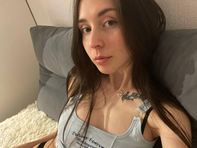 AnissaRaumhill with long brown hair, small tits in a grey tank top, black bra straps, tattoo on chest, subtle smile, lying on grey pillow, white blanket.