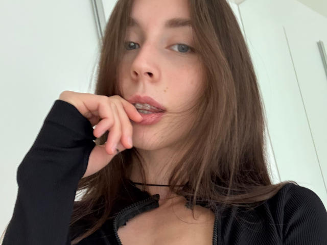 AnissaRaumhill tiene el cabello castaño, ojos azules, y está mordiéndose el dedo con los labios ligeramente entreabiertos. Lleva una blusa negra con cremallera, luciendo seductora..