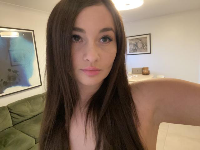 Dionisia has long brown hair, light skin, and pink lips. She's looking at the camera with a slight smile, showing her bare shoulders. Her tits are small.