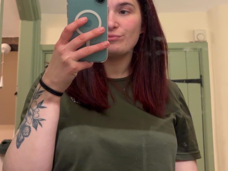 NaughtyKeeper with red hair, green shirt, holding phone, pouty face, visible tattoo on right arm, taking mirror selfie, slight blush, black wristband, in a bathroom.