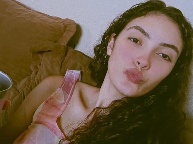 VeronyqueX has curly dark hair, pouty pink lips, and is pissing off a brown pillow while holding a cup, wearing a pink and white tank top.