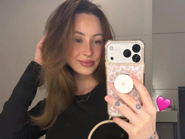 Meltattt-hot with long brown hair, wearing a black top, holding a phone with a leopard print case, smiling, freckled face, pink lips, hand in hair, pink heart emoji.