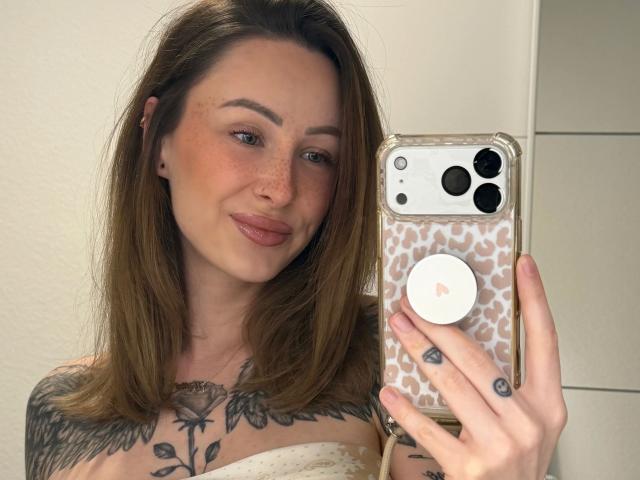 Meltattt-hot with brown hair, freckles, and tattoos, holding a phone with a leopard print case, taking a selfie, showing her tits and a rose tattoo on her chest.