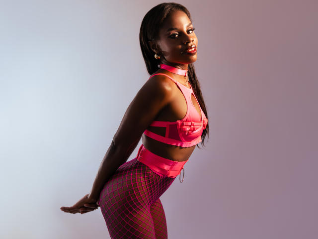 MiaHopson with dark skin, wearing pink strappy bra and fishnet pants, leaning forward, showing her ass and tits, long straight hair, colorful earrings, confident smile, sexy pose.