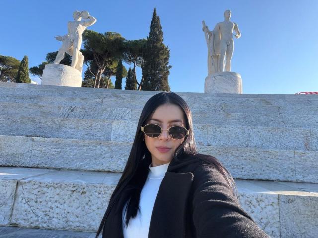 IsabellaJames with long black hair, wearing a black coat and white turtleneck, brown aviator sunglasses, standing in front of stone steps with statues, clear blue sky.