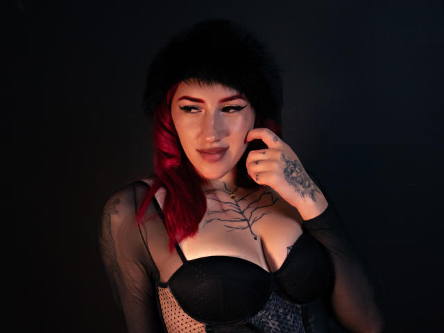 SaraJoyce with pink-dyed hair, black furry hat, black bra, sheer top, tattoos, hand near face, dark background, smiling, showing her tits, medium-sized, sexy.