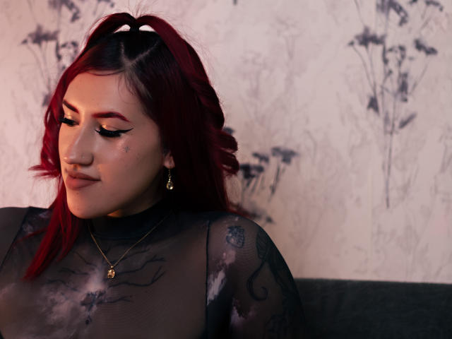 SaraJoyce with red hair, black sheer top, and gold necklace, showing off her tits, wearing bold eyeliner, and small earrings, looking down with a slight smile.