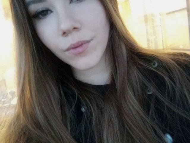 AdelMaud's face with long brown hair, pale skin, pouty pink lips, and dark eyes, wearing a black shirt. She's looking slightly to the side with a subtle smile.