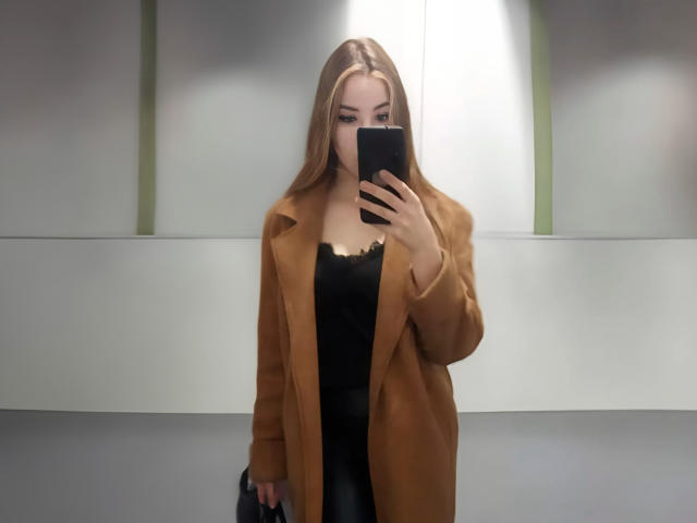AdelMaud with long blonde hair, wearing a brown coat over a black top, holding a phone in front of her face, standing with a black bag in her other hand.