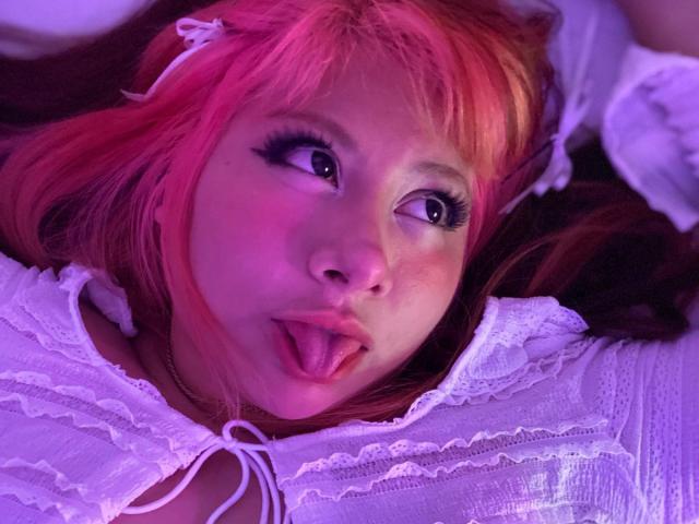 AngelineWoods with pink hair, sticking out her tongue, wearing a white lace top, has purple and pink lighting on her face, looking up with big eyes and long lashes.
