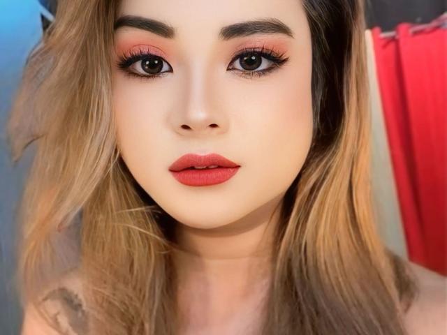 Close-up portrait of AlluringJhana with fair skin and long, wavy blonde hair. She has bold, dark eyebrows, prominent eyeliner, and pink eyeshadow. Her lips are painted a deep red. The image focuses on her face, with a slight blur on the background. The lighting is soft, highlighting her facial features. Her expression is neutral, with a slight upward tilt of her head. The texture of her hair is smooth, and her makeup is meticulously applied.