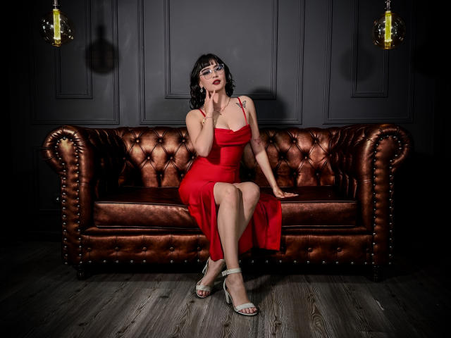 ElaJamess sits on a brown leather couch, wearing a red dress that shows her tits, silver heels, and glasses, with one hand on her face, looking sexy.