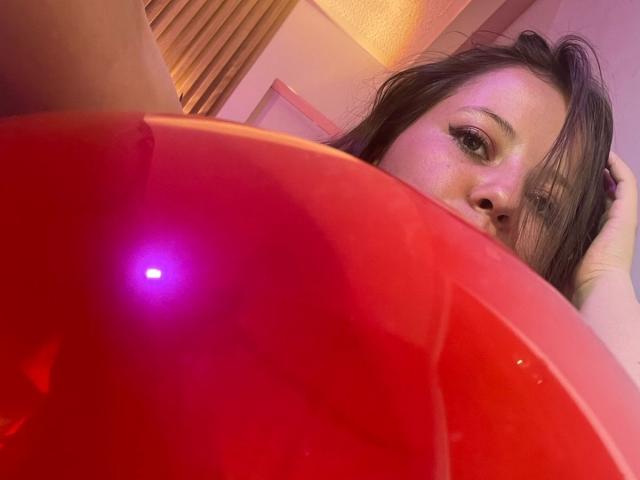 GraceRedMiller with long brown hair, looking up from behind a shiny red plastic surface with a purple light reflection, her face partially visible, in a pink-lit room.