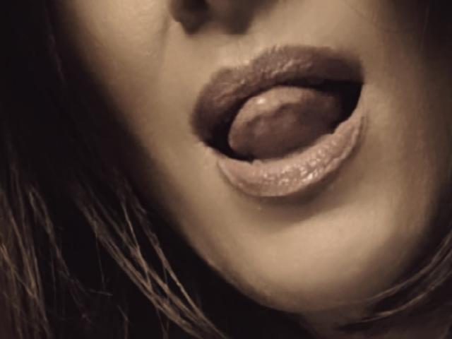 Close-up of SophieDesir's face with a sensual, slightly open mouth. Her tongue is partially out, touching her upper lip. She has full, glossy, dark brown lips with a subtle sheen. The image has a sepia tone, giving it a vintage feel. Her skin is smooth with a slight shadow on her chin. Her dark hair is slightly wet, hanging over the right side of her face. The focus is on her lips and tongue, with no other visible details. The texture of her lips is slightly glossy and textured.
