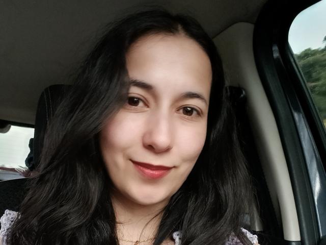 AnneStone a la peau pâle, les yeux marron et les cheveux noirs ondulés, avec un léger sourire et des lèvres rouges, assise dans une voiture. Ses seins sont petits, et elle porte un haut en dentelle..