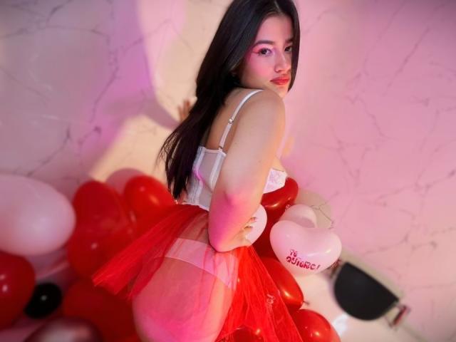 Iris Blair, con lunghi capelli scuri, reggiseno bianco e gonna di tulle rossa, seduta su palloncini rossi e bianchi, che si guarda indietro con un'espressione seducente, mostrando il sedere..