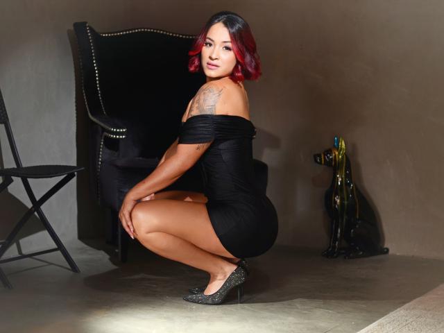 ValeryMonnroy, squatting, wearing a black off-shoulder dress, showing a tattoo on her left shoulder, with red hair, black heels, and a confident smile.