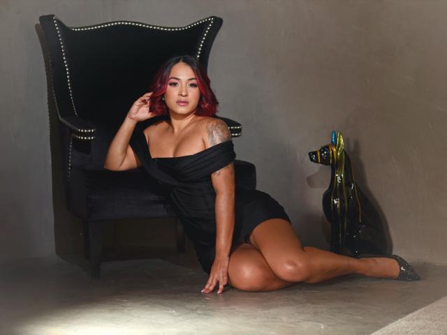 ValeryMonnroy with red hair, black off-shoulder dress, large tits, tattoo on right arm, sitting on floor, black chair with gold studs, black heels, and black sculpture in background.