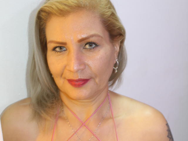 VictoriaRubio has blonde hair, blue eyes, red lipstick, and a slight smile. She's wearing a pink halter top with silver earrings. Her skin is slightly shiny.