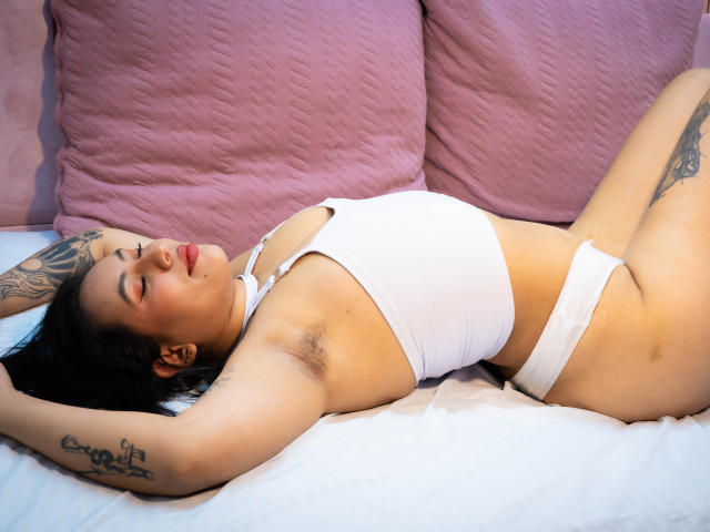 Amy Ross está acostada en una cama, vestida con un bikini blanco, con los brazos por encima de la cabeza, mostrando vello axilar, tatuajes y una expresión relajada. Tiene los ojos cerrados y está sobre almohadas rosas..