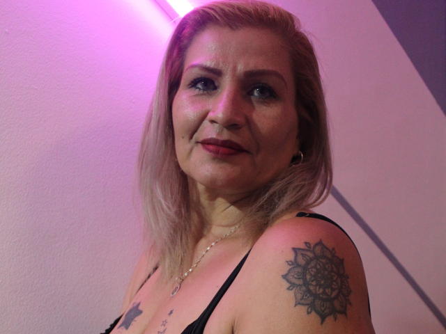 VictoriaRubio has blonde hair, blue eyes, and a slight smile. She has tattoos on her right shoulder and wears a black strap top with a necklace. Pink lighting.