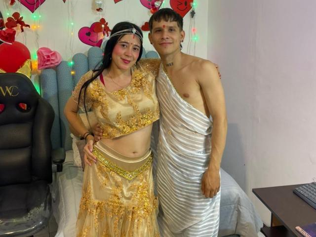AnjaliRohan, a woman with medium skin and long black hair, wears a gold-embroidered, sheer beige outfit with a headpiece. She stands next to a man with light skin and short brown hair, wrapped in a white, one-shoulder toga. The man's chest is partially exposed. Both are smiling. Decorative heart-shaped and colorful wall decorations are in the background. The man's toga is shiny, contrasting with AnjaliRohan's delicate, ornate outfit. The image has a casual, festive vibe.