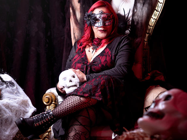Sammy Owens, con i capelli rossi, una maschera di pizzo nero, un abito rosso e nero, calze a rete, stivali neri, che tiene una maschera bianca, seduta su una sedia elaborata, circondata da maschere..