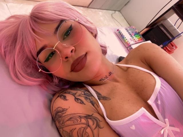 LenaBranch with pink hair, green eyeshadow, and round glasses, lying on a bed, wearing a pink and white bralette, showing tattoos on her left shoulder and chest.