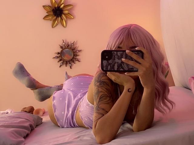 LenaBranch met roze haar, liggend op een bed, gekleed in paarse shorts met stippen, een wit tanktopje, grijze sokken, een getatoeëerde arm, vasthoudend een telefoon, een spiegelzelfportret makend, waarbij haar achterwerk te zien is..
