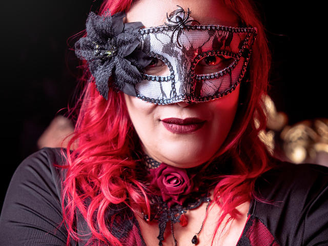 SammyOwens ha i capelli rossi, l'eyeliner nero e una maschera di pizzo con un fiore nero, indossa un trucco scuro e una collana con una rosa nera. Il seno è visibile sotto la maglia di rete nera..