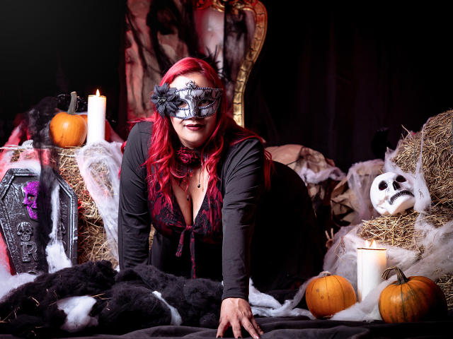 SammyOwens with red hair, black mask, and cleavage, on hands and knees, surrounded by candles, pumpkins, and black fur, in a spooky Halloween setup.
